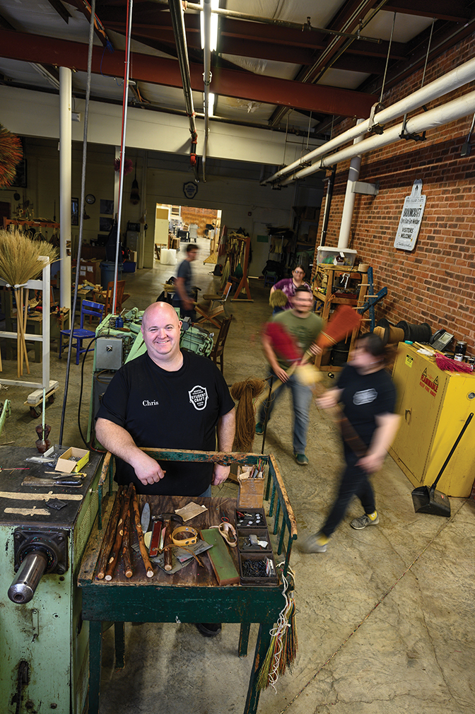 Broommaker Christopher Robbins stands in Student Craft while busy students move around him in blurred motion
