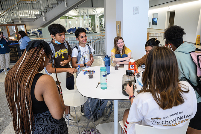 Students gathered around a table talking in the MAC building lobby. 