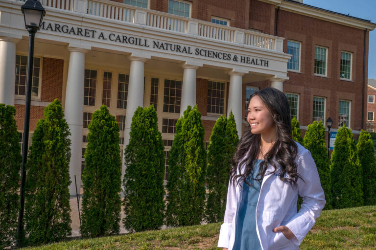 Kimberly '26 stands in her lab coat in front of the MAC building