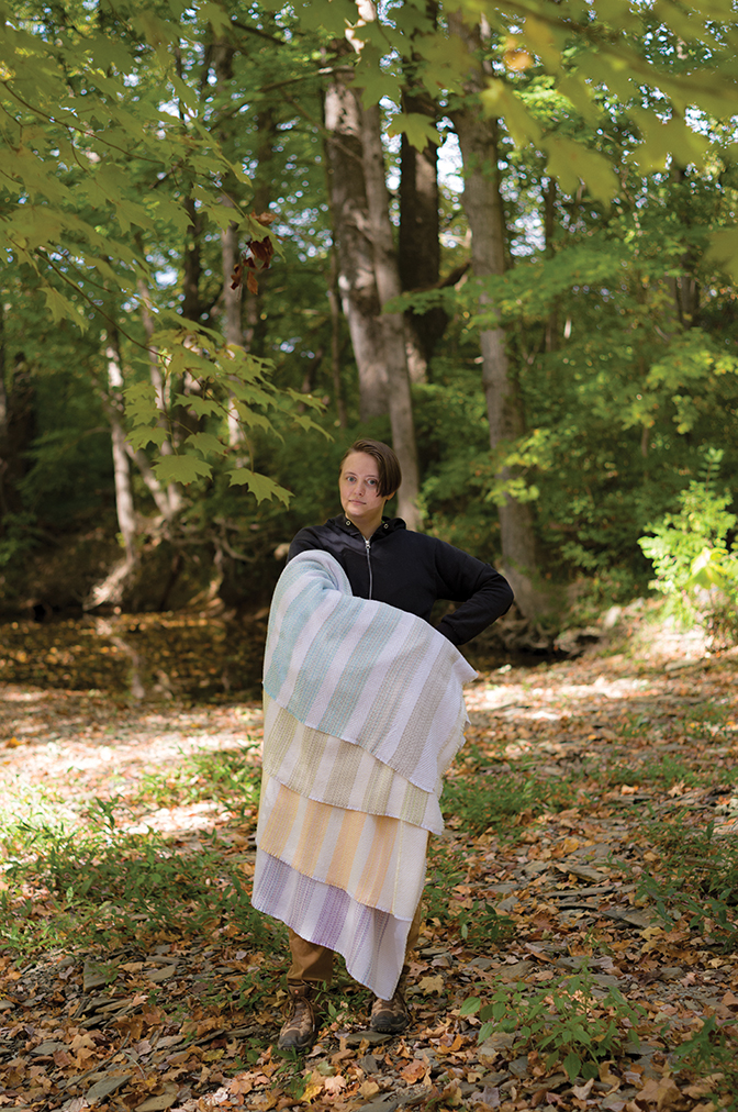 Emerson Croft '21 stands in the woods with a blanket they designed while a student in Berea College Craft