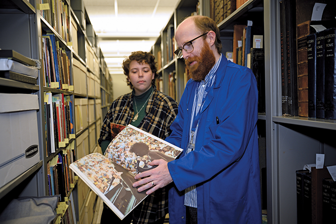 Peter Morphew and Alivia West stand amid the Berea College archive shelves looking at a magazine with an old image of Janis Ian at a concert