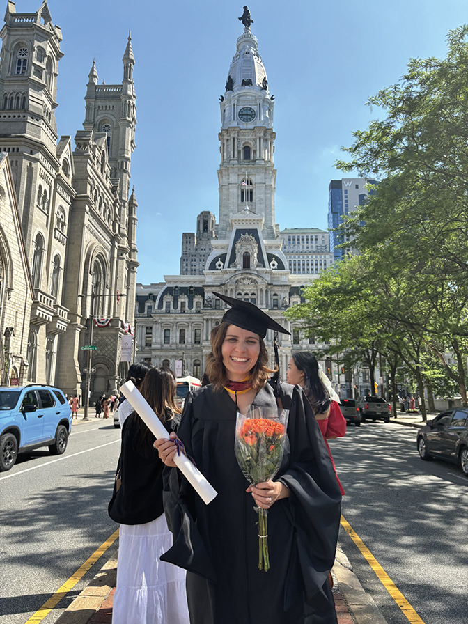 Photo of Rachel Mauro Hutchins standing in a downtown area in graduation cap and gown, holding flowers