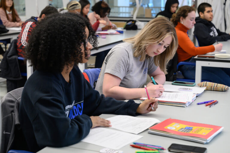 Two female presenting students sitting at a table in taking notes in a science classroom in the MAC building