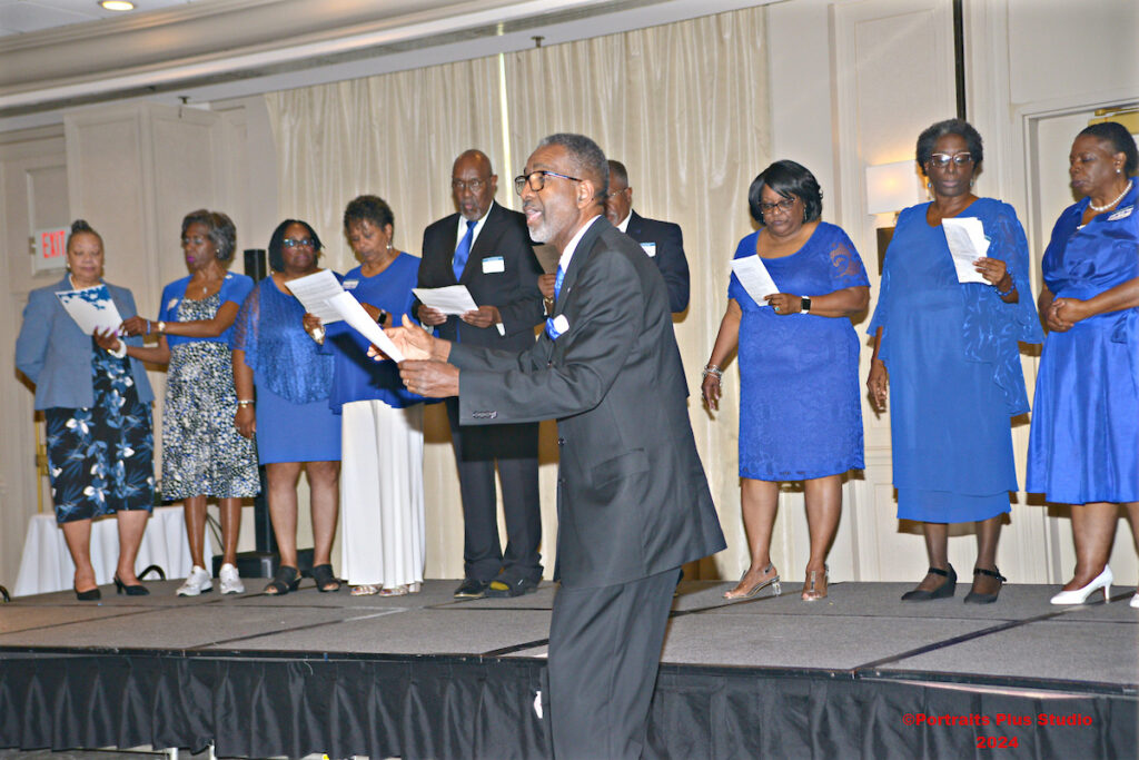 Choir director leads gathered alumni in songs with a choir behind him