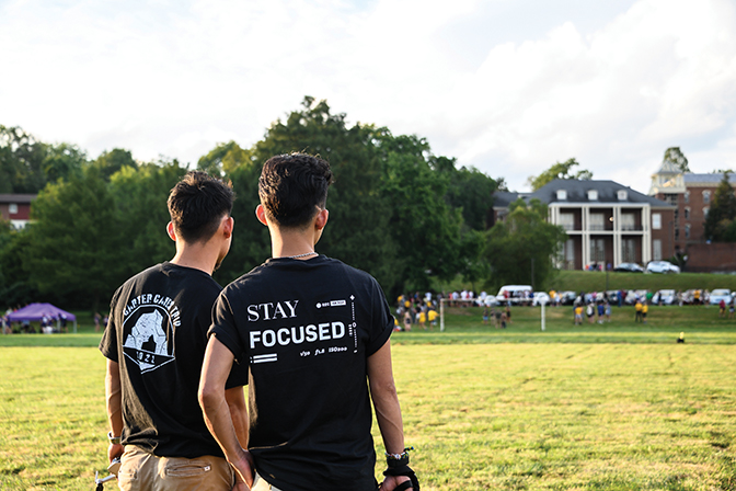 Photo of two young men standing with their back to the camera on Alumni Field at Berea College. They are wearing Berea College photography team shirts.