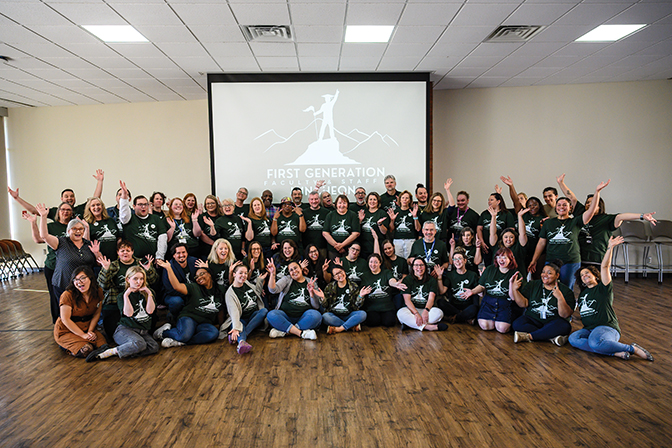 A large group of first-generation staff and faculty pose on a wooden floor.