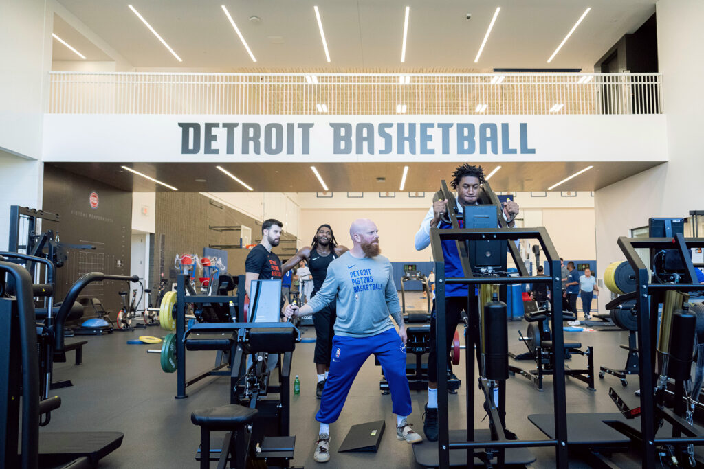 Cory Schlesinger ’09 stands beside an exercise machine in the Detroit Pistons Basketball training facility helping a player reach his full potential on the machine. 