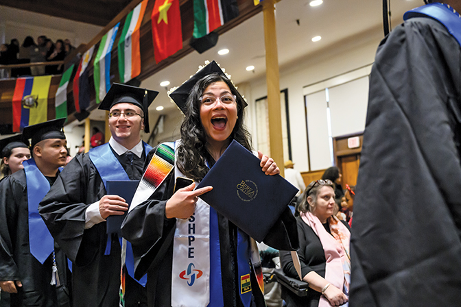 A student in graduation regalia points at her diploma with a line of graduates behind her.
