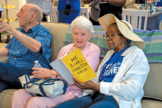 Two women sit on a couch together reading a pamphlet on the history of the Best-Evans house