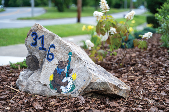 Image of a rock painted with 316 and a bear playing a banjo. The rock sits in a flower bed in front of the Best-Evans house.