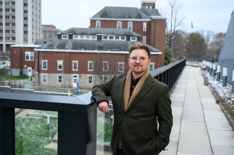 We Are Here Holden Dillman standing on a balcony outside with a brick building in the background.