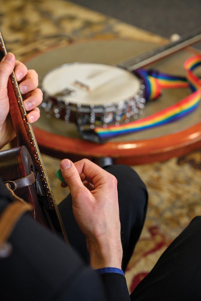 Upclose photo of hands picking at a banjo neck, with a banjo in the background with a raninbow neck strap.