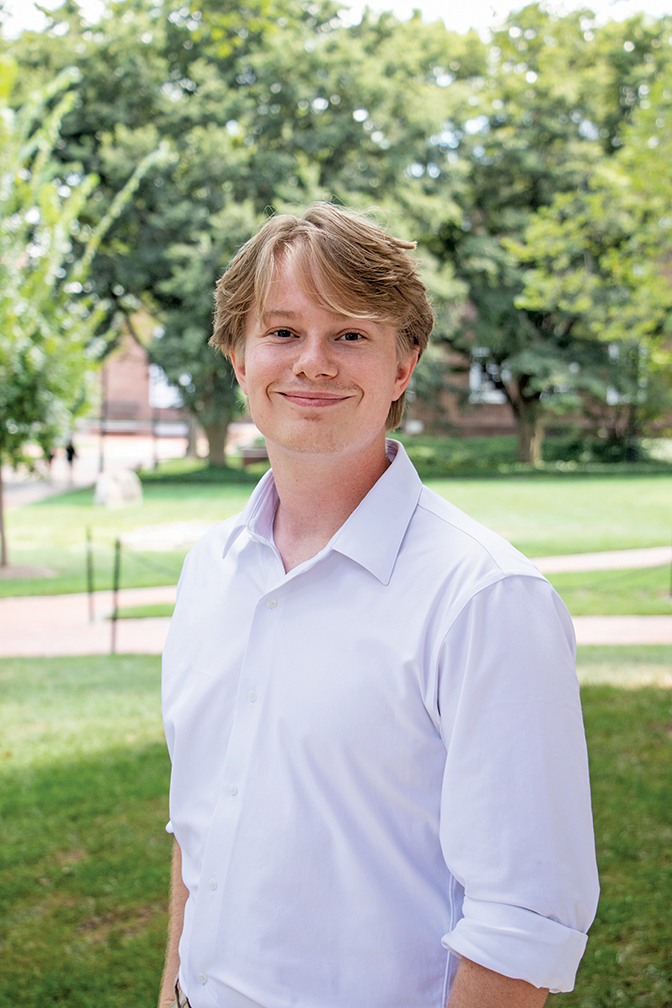 William Woelki is wearing a light-colored collared shirt. He is standing outside on a green campus with trees behind him.