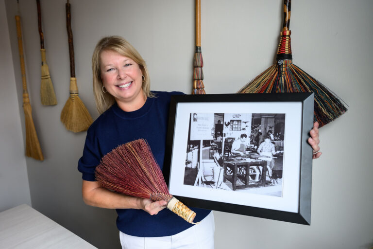 Deloris Reed poses in her office with a black-and-white photo of her parents working on brooms. She also holds a broom that her father made that was gifted back to the family.
